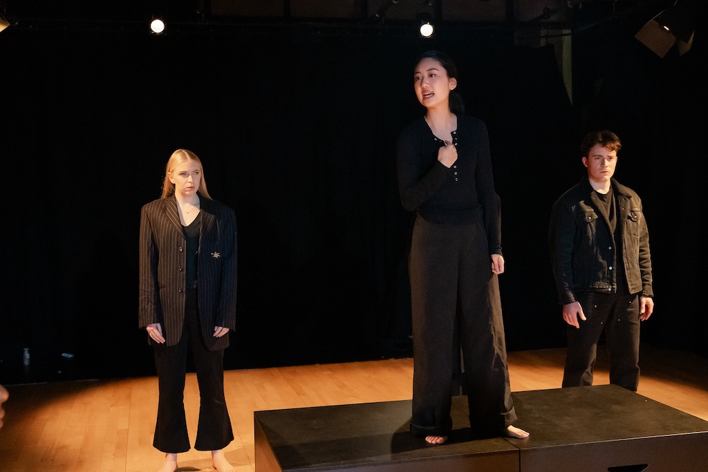 An actor stands on a black riser in mid-speech, while two other actors stand behind her.